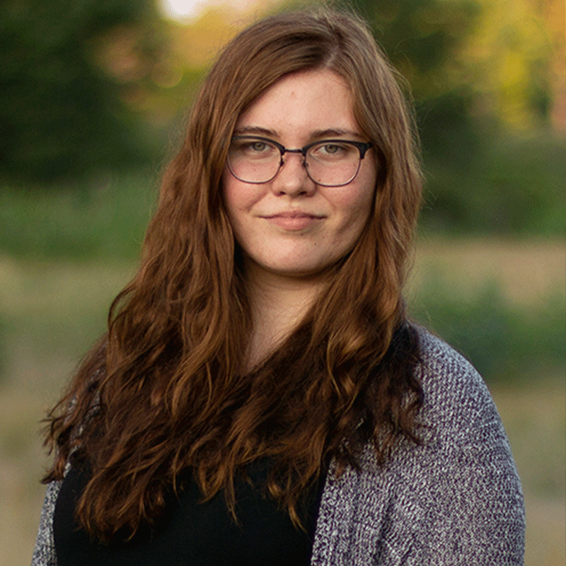 Image of a woman with long chestnut hair and glasses wearing a grey sweater over a black shirt