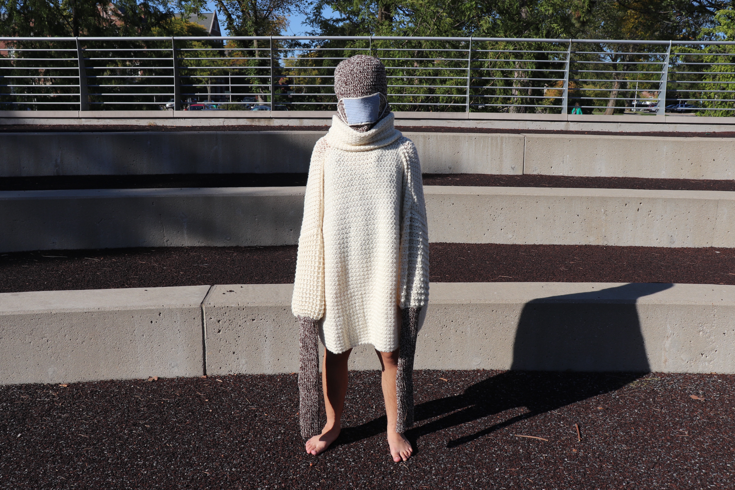 A person standing dressed in a white long knitted turtleneck with another sweater underneath covering all of the head and stretching past the arms. A blue disposable mask is worn on nose and mouth area.