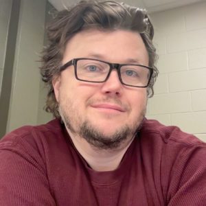 Man with a beard, glasses, and a red shirt smiling at the camera.