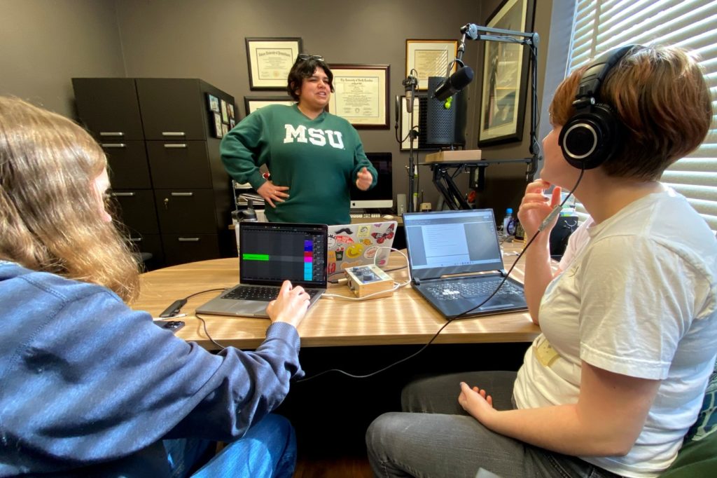 A photo with three people, two who are working on computers, one of whom is wearing headphones, and the third person is standing at a microphone. 