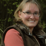 Imari, a white woman with short light brown hair and glasses, is smiling in front of a leafy background.