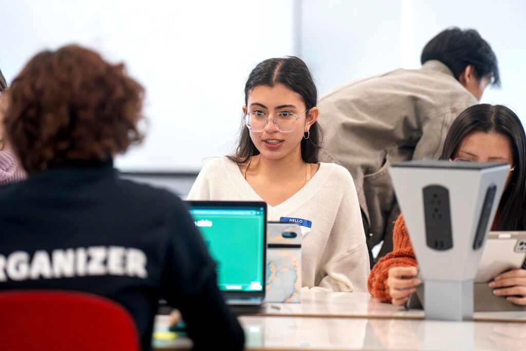 A woman with long black hair that is pulled back and is wearing glasses and a white v-kneck sweater is sitting at a table with a computer and it looking across at another person with computer.