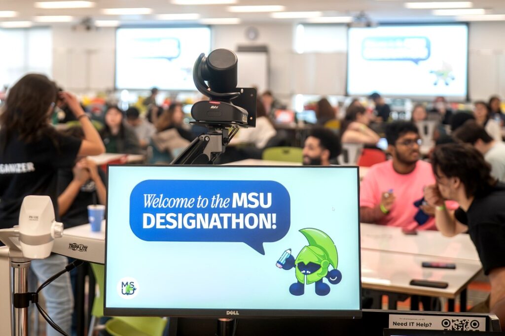 A digital display in the foreground reads “Welcome to the MSU Designathon!” alongside a cartoon character in a green helmet holding a pencil. A video camera is mounted above the screen, and a large group of students is visible in the background