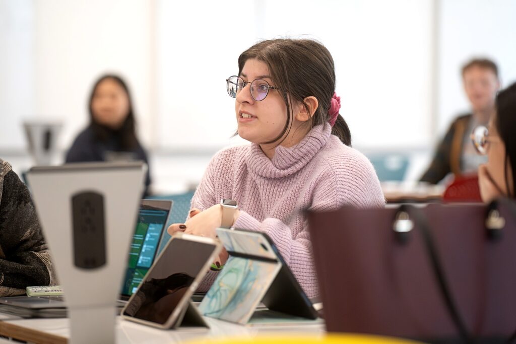 A student in a lavender sweater speaks during a group discussion at a table surrounded by tablets and laptops. She wears round glasses and has a pink scrunchie in her hair. Other participants are visible in the background, slightly out of focus.