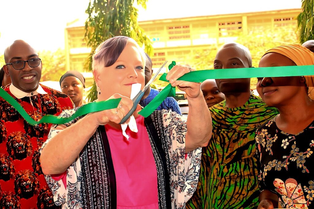A woman with short platinum-blonde hair holding scissors and doing a ribbon cutting. She is wearing a bright pink top top with a white and black patterned jacket on top. Behind and beside her are people watching her cut the ribbon. 