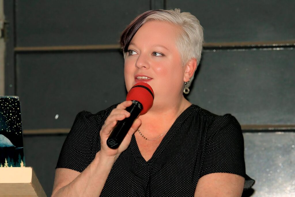 A woman with short platinum-blonde hair holds a red microphone and speaks at a podium. She is wearing a black top with white polka dots and small hoop earrings. A dark background and part of a laptop with a starry night design are visible beside her.