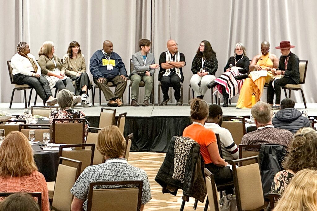 A diverse group of speakers sit in a row on stage, engaged in discussion during a panel session at the Conference on Community Writing. Audience members watch from tables in the foreground.