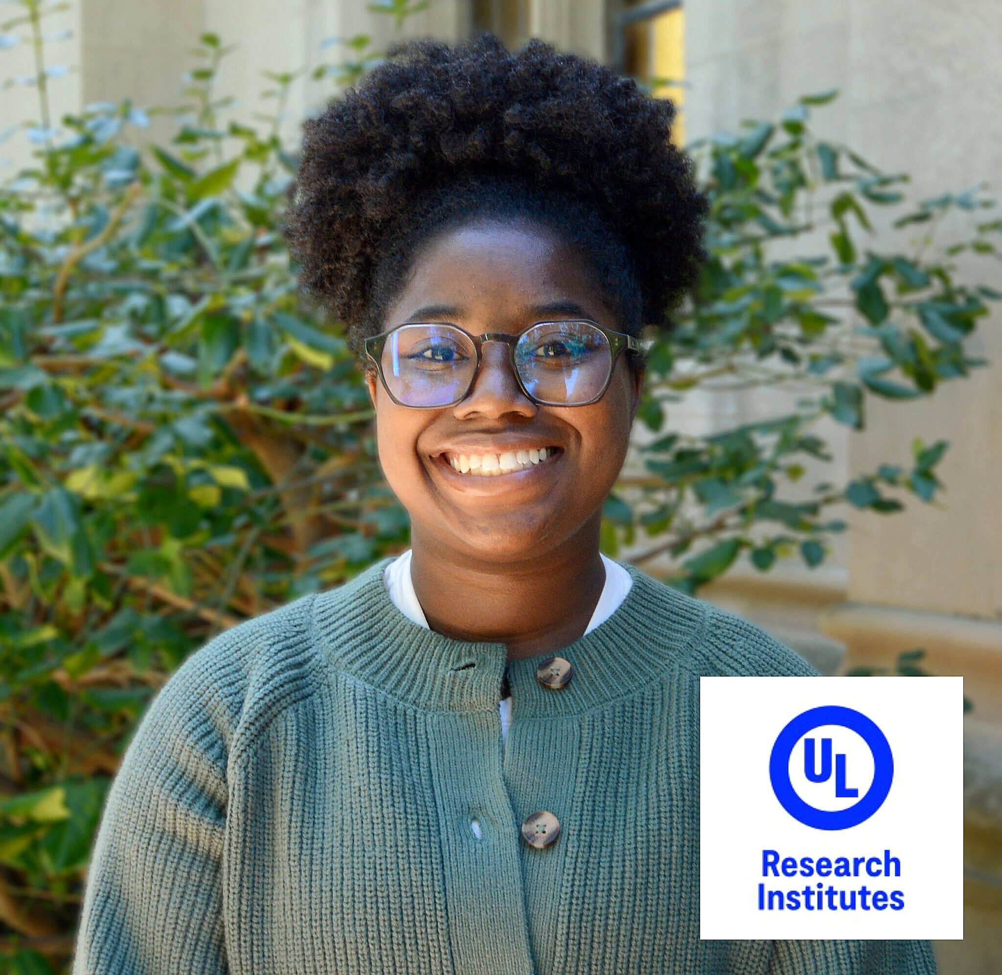 A person wearing glasses and a green sweater smiles in an outdoor setting with greenery and a building in the background. The UL Research Institutes logo appears in the lower right corner.