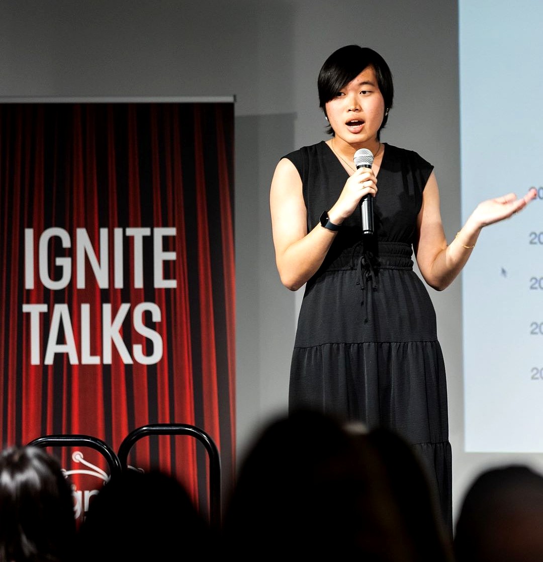 An Asian woman with black short hair and an all-black sleeveless dress with a v-neck, holding a silver microphone and wearing a black watch. She is on a stage with a banner display behind her that says "Ignite Talks" in white letters on a red background.
