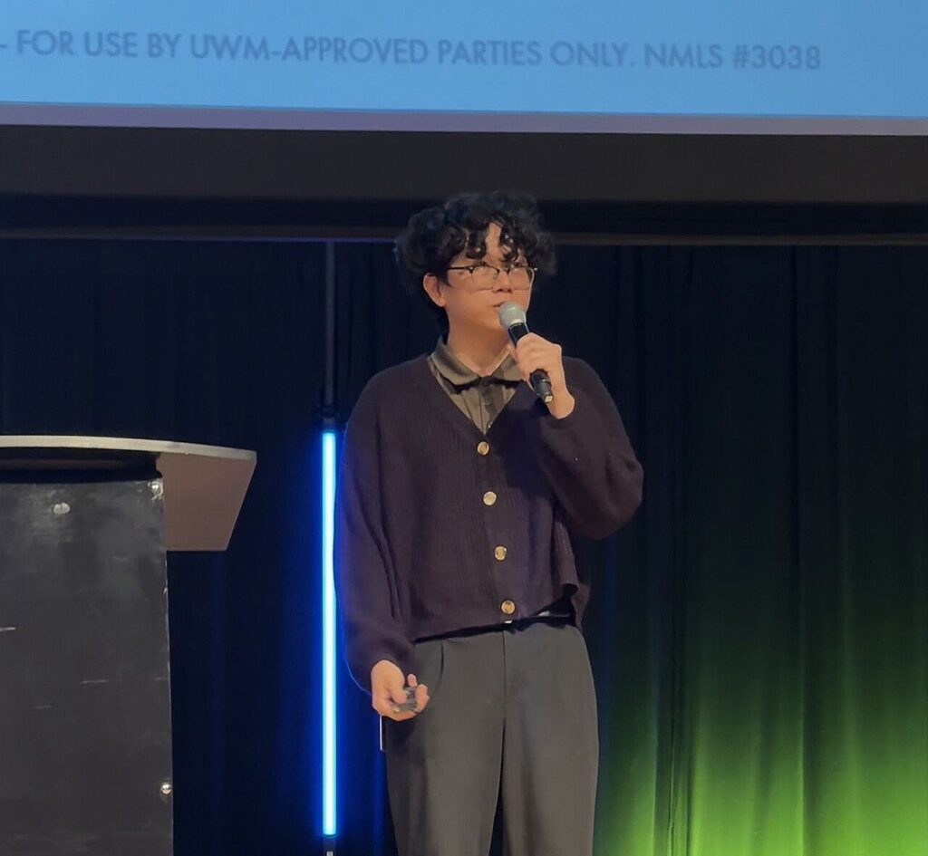 Man, dressed all in black with short black curly hair and glasses, holding and speaking into a microphone on a stage with a dark background.