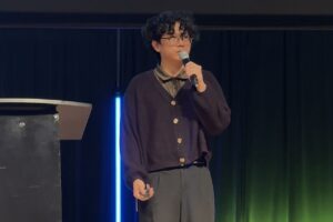 Man, dressed all in black with short black curly hair and glasses, holding and speaking into a microphone on a stage with a dark background.