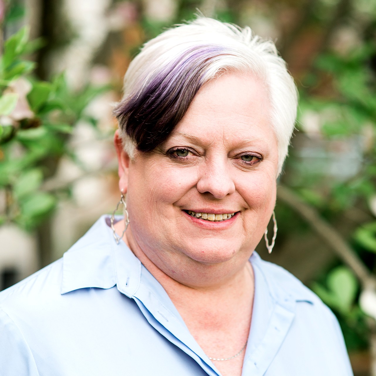 A close-up outdoor portrait of a person with short, white hair with a purple streak, wearing dangling earrings and a light-colored shirt, smiling at the camera with greenery in the background.