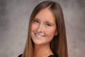 A professional headshot of Avery Kuehl. She has long, straight brown hair, blue eyes, and is smiling while wearing a black top and a gold chain necklace against a textured gray background.