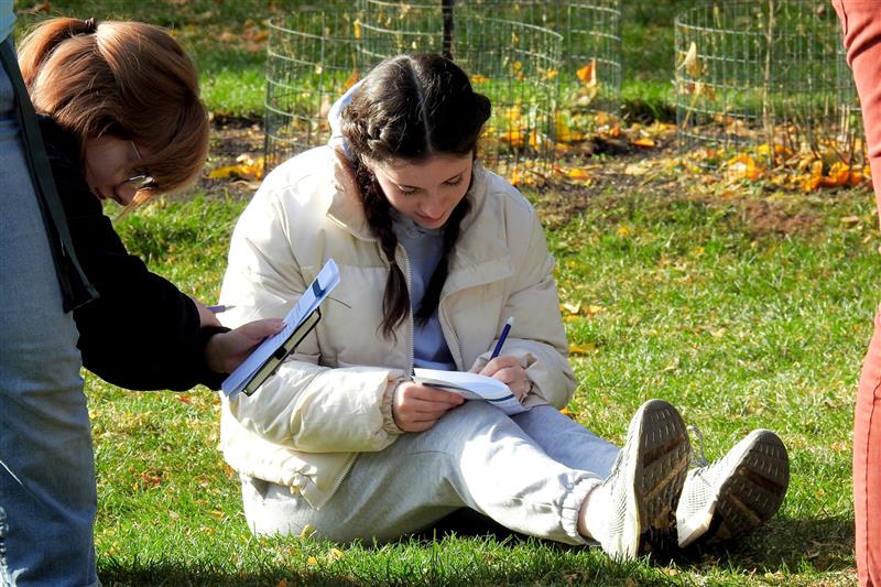 An Experience Architecture student sits on the grass at Beal Botanical Garden, focused on writing in a notebook. Another student stands nearby. 