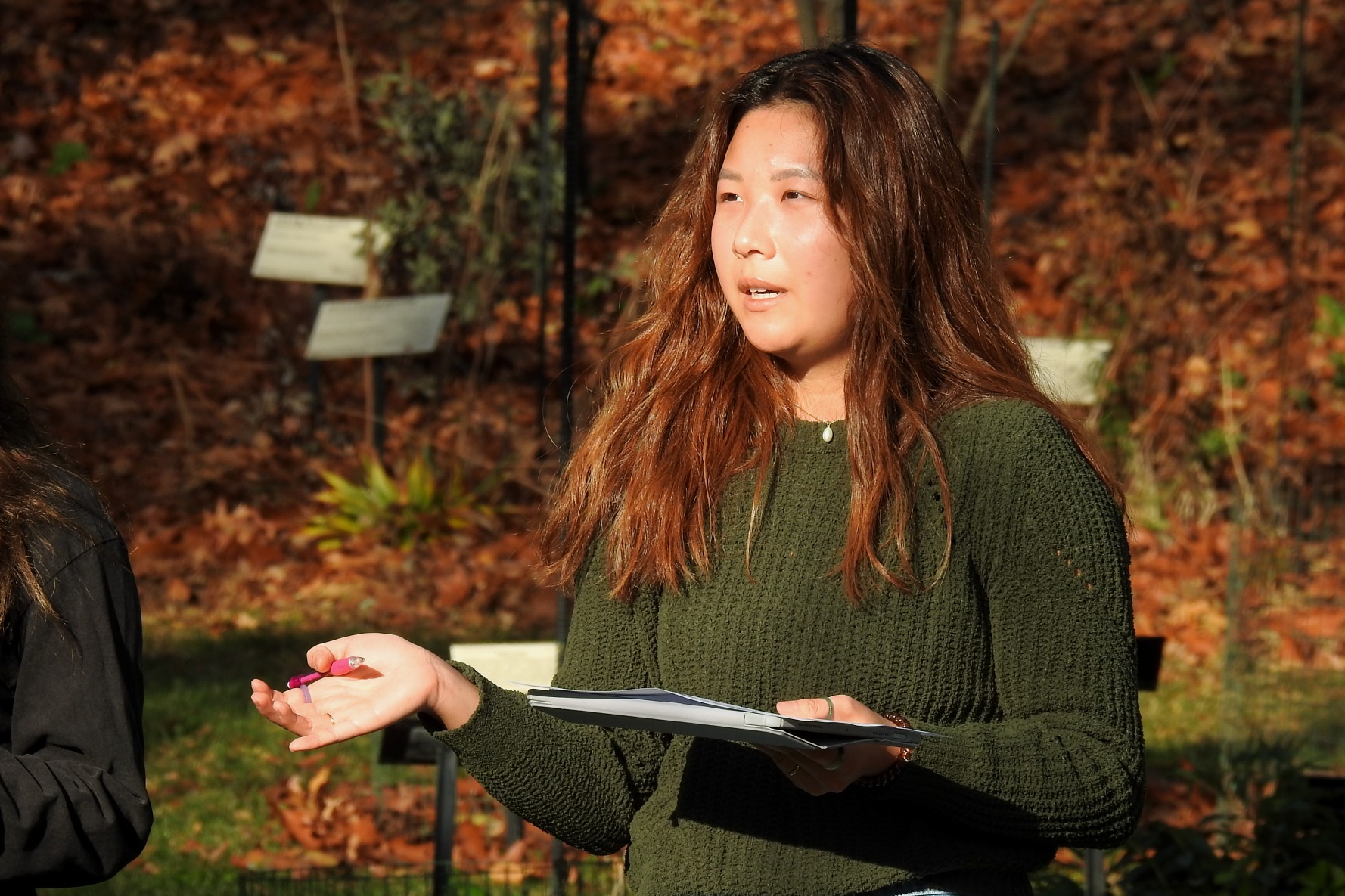 An woman in a green sweater stands in Beal Botanical Garden, holding a notebook and gesturing with a pen. 