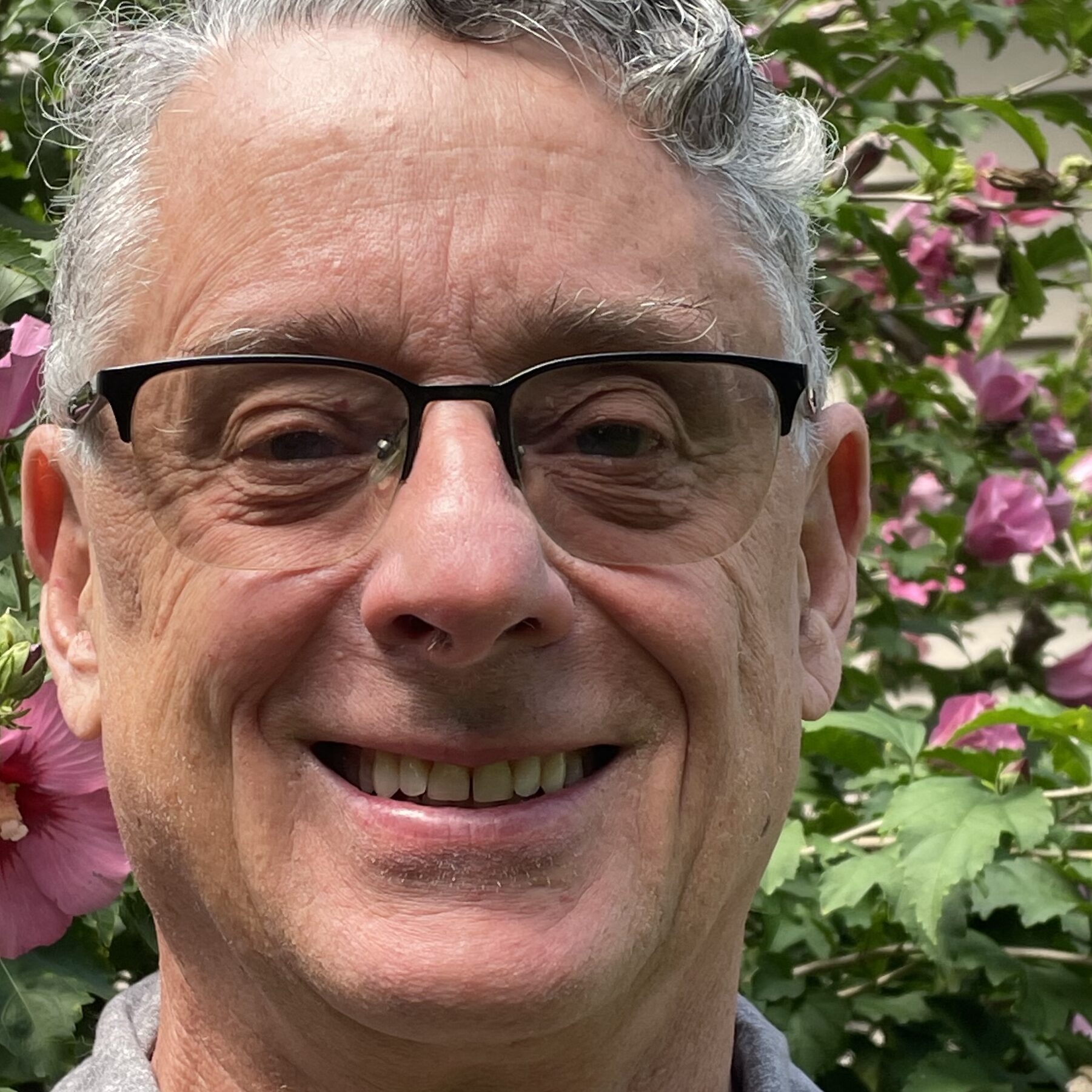 Close-up headshot of Matt McKeon with graying hair and black-rimmed glasses. He is smiling broadly in front of a natural background of green leaves and large pink hibiscus flowers.