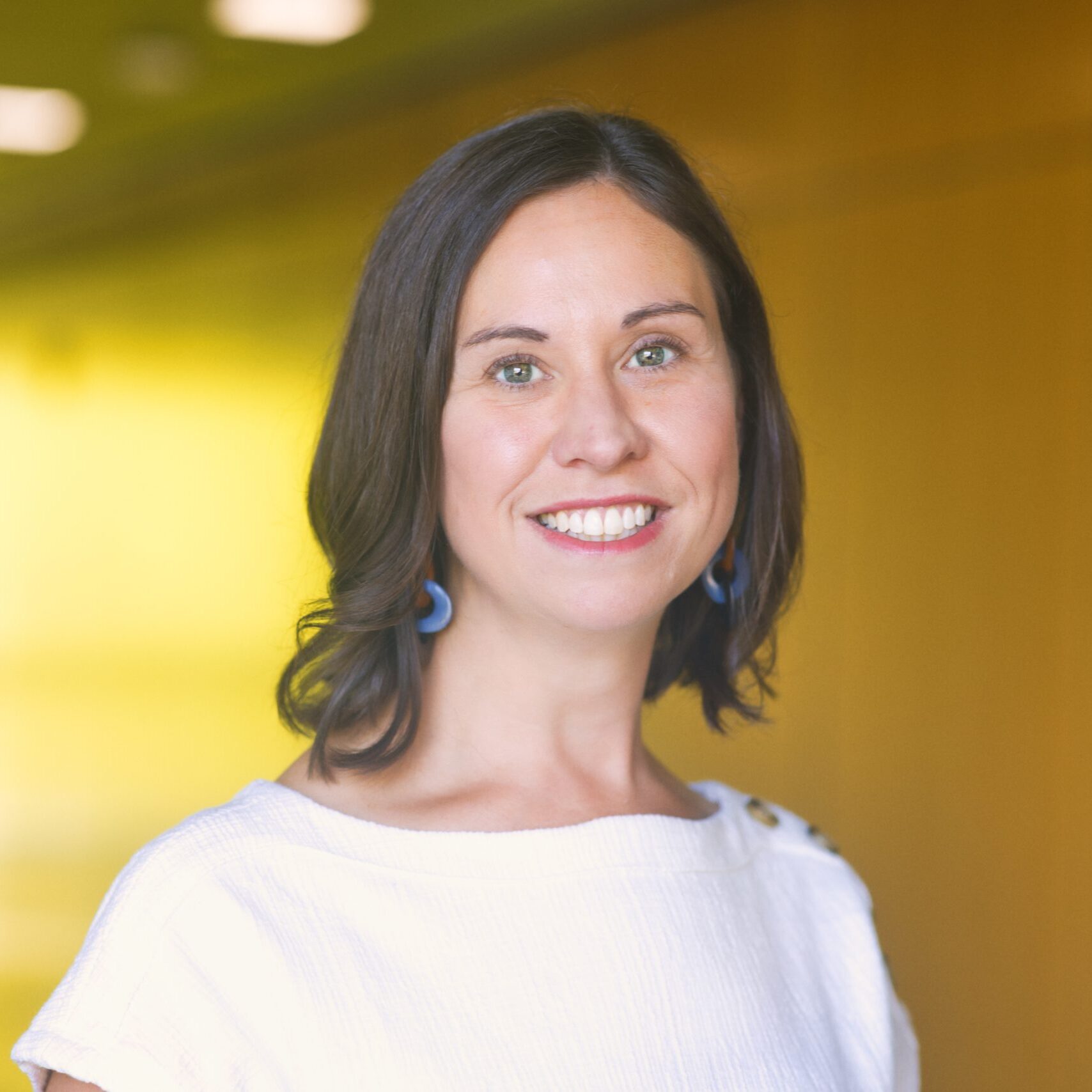 Professional headshot of Laura Yares smiling and wearing a white boat-neck top with gold button details on the shoulder and blue hoop earrings. The background is a warm, blurred yellow and orange.