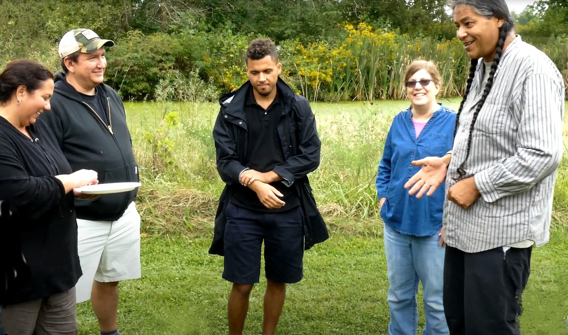 Five people standing outdoors in a grassy field with trees and yellow wildflowers in the background. The group includes individuals wearing a white baseball cap and black jacket, a black jacket, a blue button-up shirt, light-colored pants, and a gray striped button-up shirt.