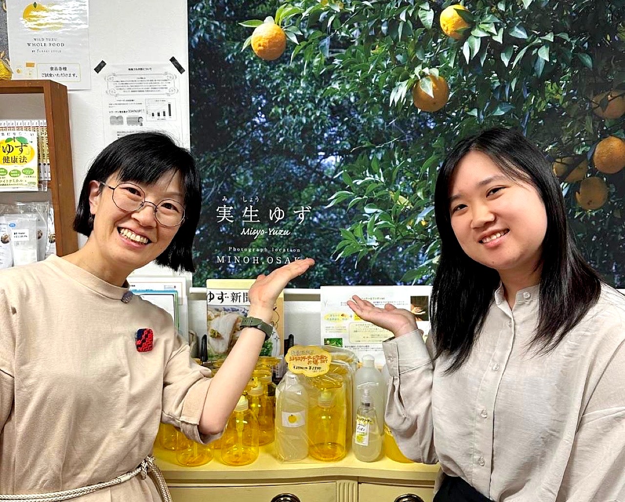 Two women stand indoors, smiling and gesturing toward a display behind them featuring a large photo of citrus fruit growing on a tree, with shelves of yellow bottles and packaged goods arranged below the poster.