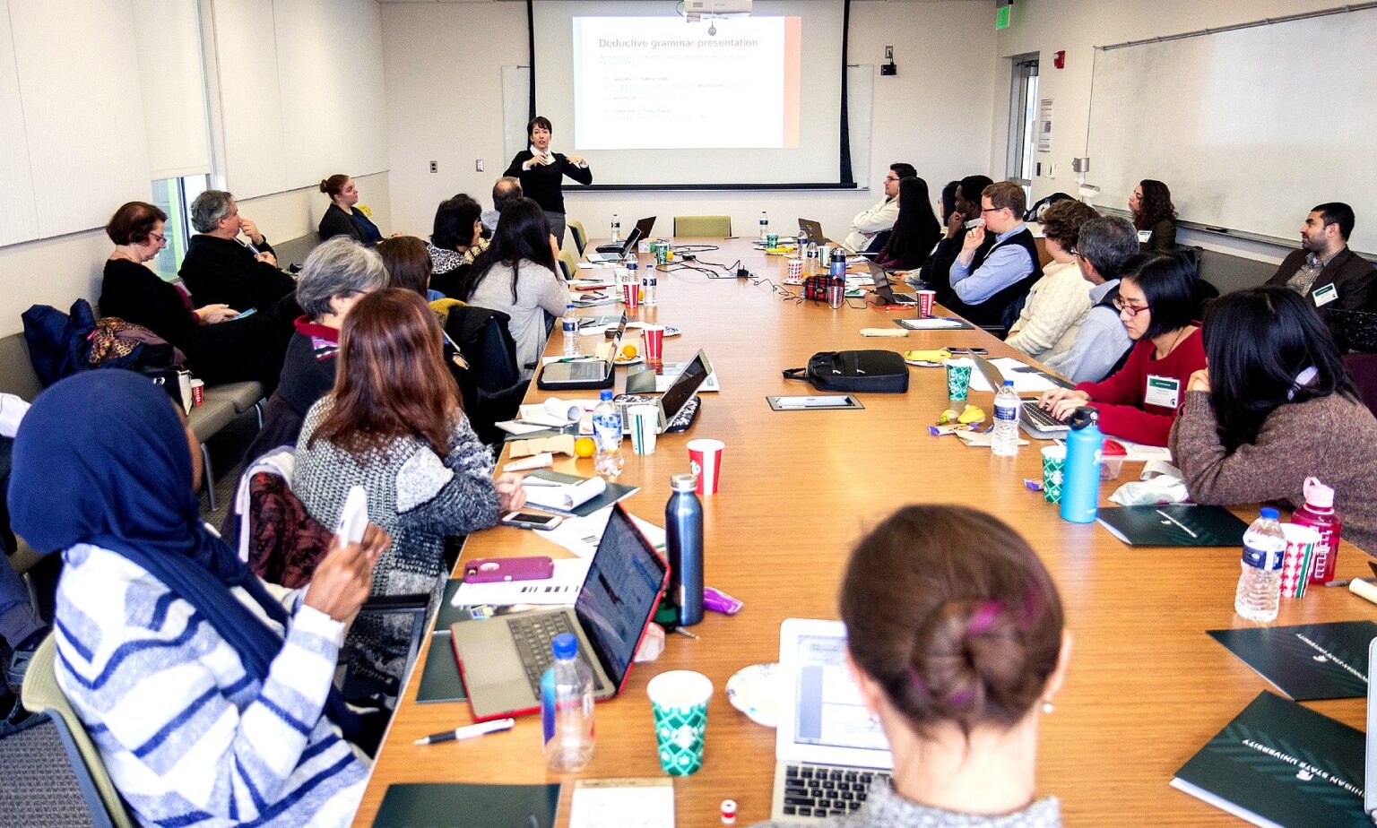 A large group of language instructors from across the Big Ten Academic Alliance attend a professional development workshop, seated around conference tables in a classroom with a presenter speaking at the front.