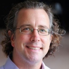 Max Evjen, wearing glasses and a light purple button-up shirt, smiling at the camera against a neutral gray background.