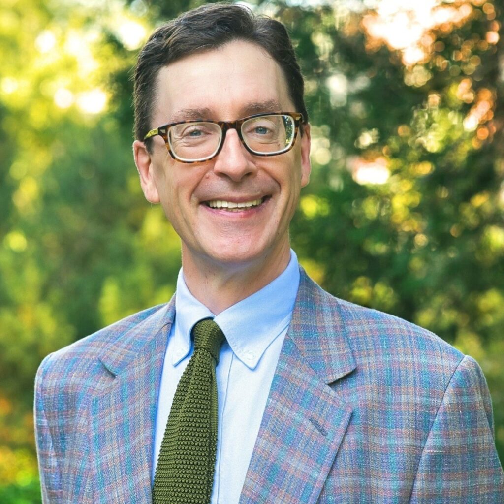 Stokes Schwartz, wearing glasses, a blue shirt, and a green knit tie with a blue and green plaid sport jacket, smiling at the camera against a green foliage background.