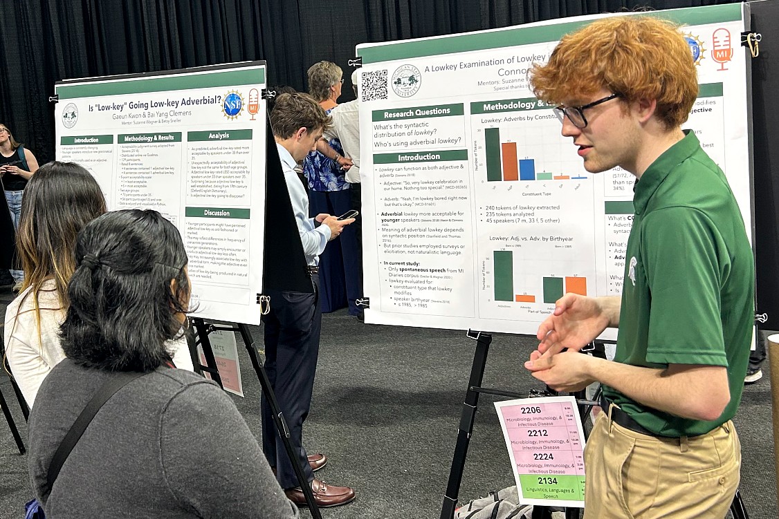 A young man discusses research at a poster session. He's wearing a green shirt and glasses, passionately explaining charts on a board. Attendees listen attentively.