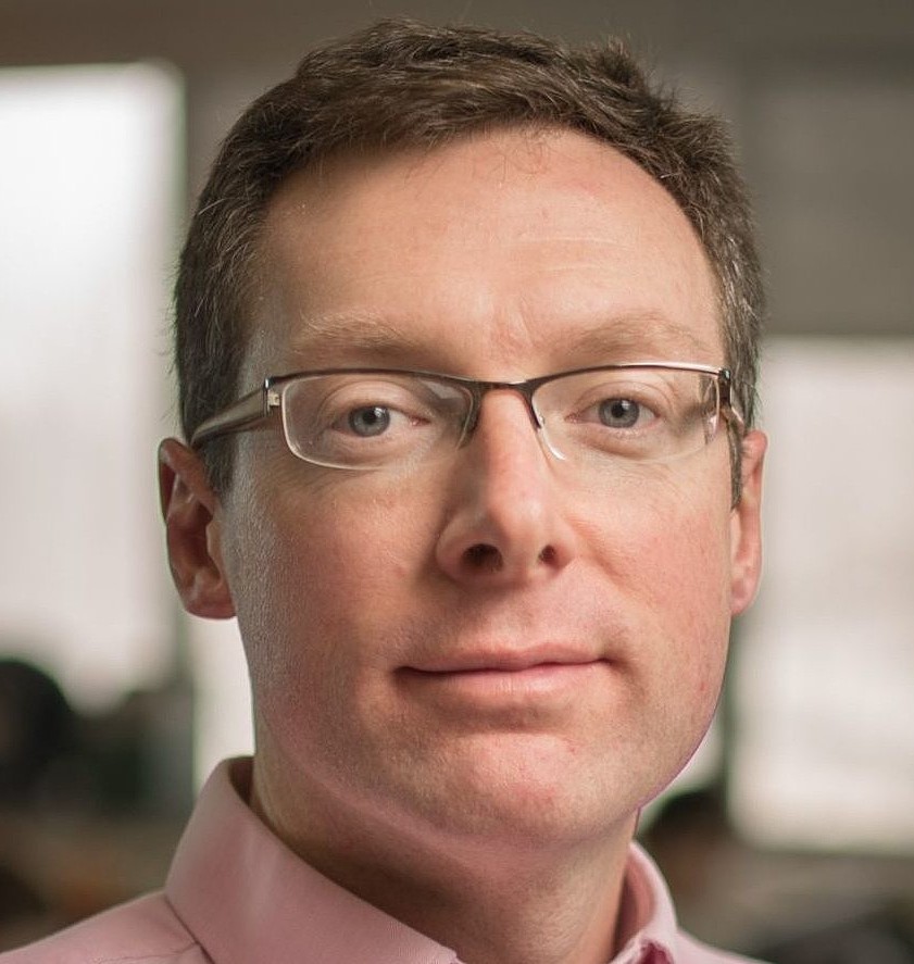 A man with glasses and short brown hair in a pink shirt appears focused, gazing at the camera. Soft lighting and blurred background convey a calm, professional setting.