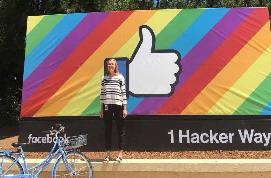 smiling woman in front of a rainbow striped sign with the facebook "like" symbol on it