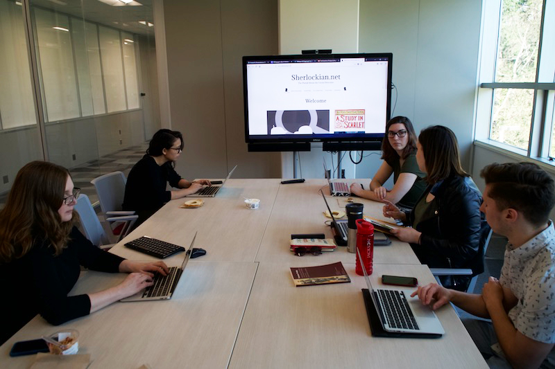 Group of people at a conference table working on laptops while a project is displayed on a tv behind them
