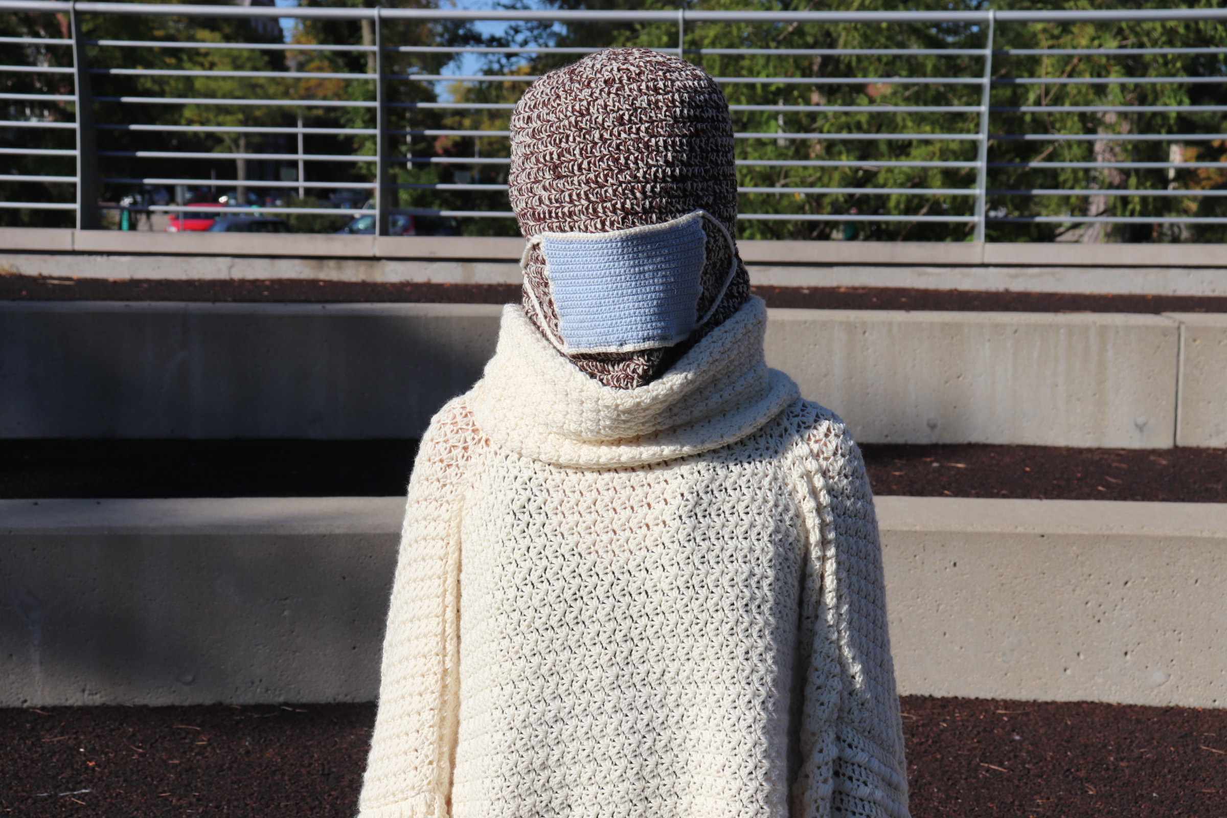 Photo of a zoomed in view of person wearing an oversized white knitted turtleneck with a different sweater covering the entire head and a disposable blue mask put on top of the face.