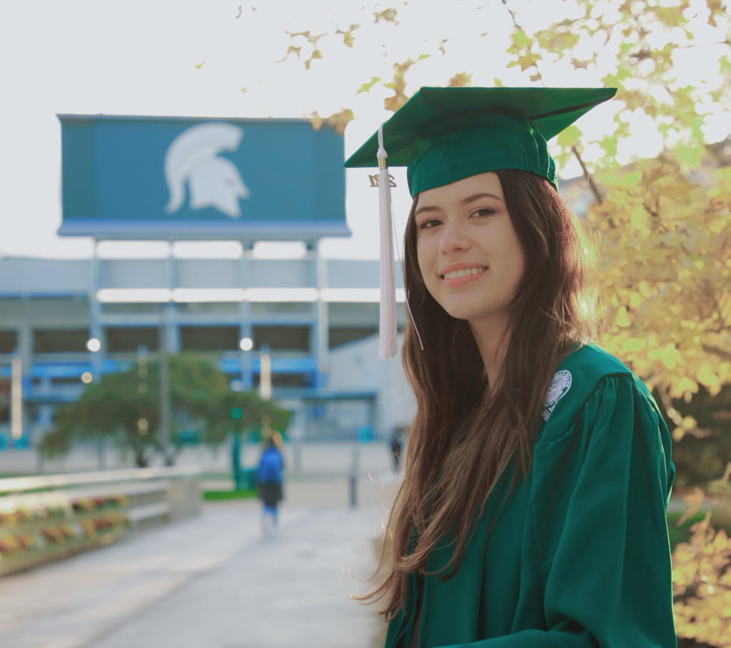 Woman with long brown hair wearing a green graduation cap and gown. 