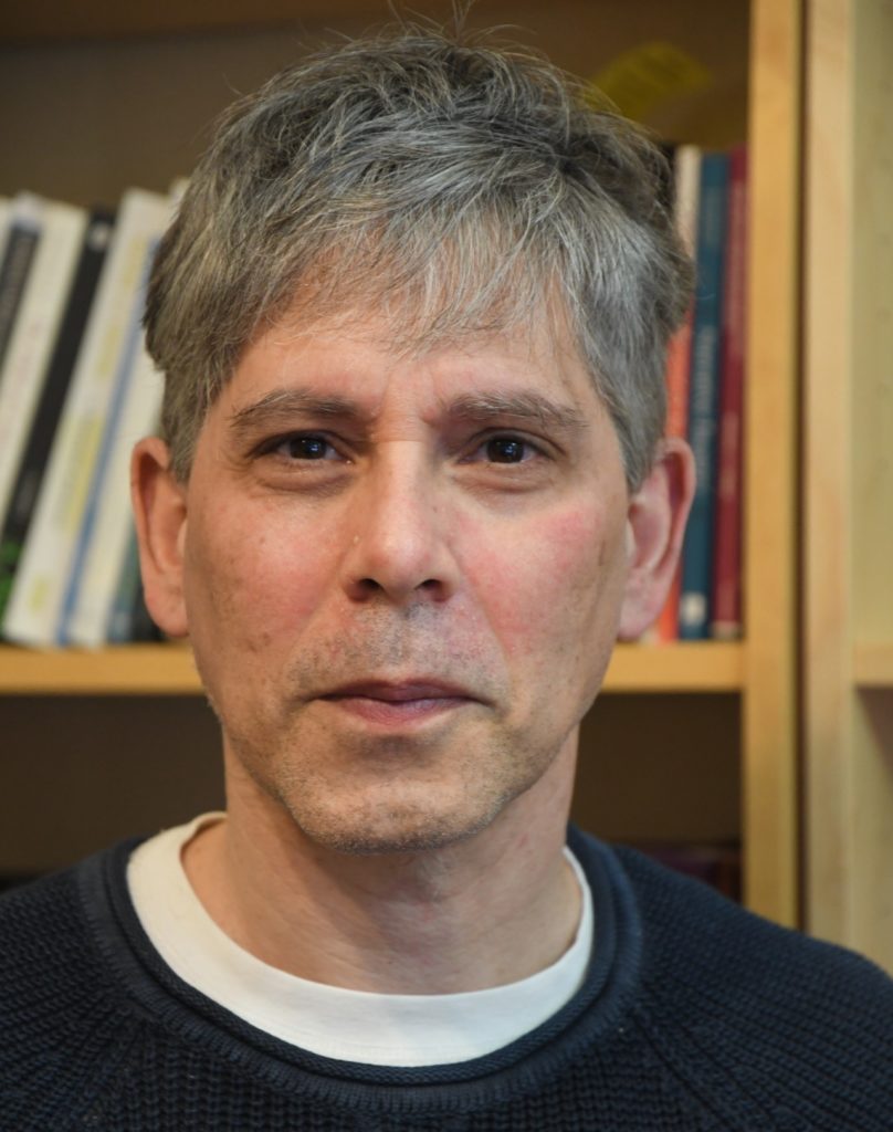 Headshot of a man with gray hair in a navy blue sweater standing in front of a bookshelf.