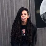 Woman with black dreadlocks and a black shirt smiling in front of a gray wall.