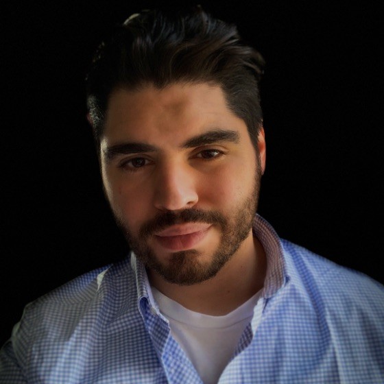 Headshot of a man with black hair and a beard wearing a blue and white button up shirt