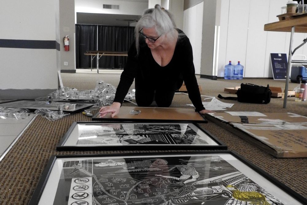 A woman with glasses and gray hair wearing a black top and pants kneels on the floor as she fits art work into frames. More framed art works and other materials surround her.