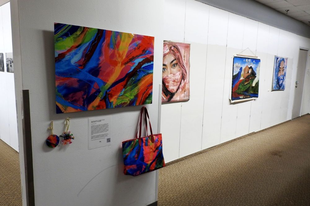 Bright works of art hang on the wall of an exhibit. Underneath an abstract painting hangs a handbag of the same pattern with three paintings of women wearing COVID-19 face masks further to the right.
