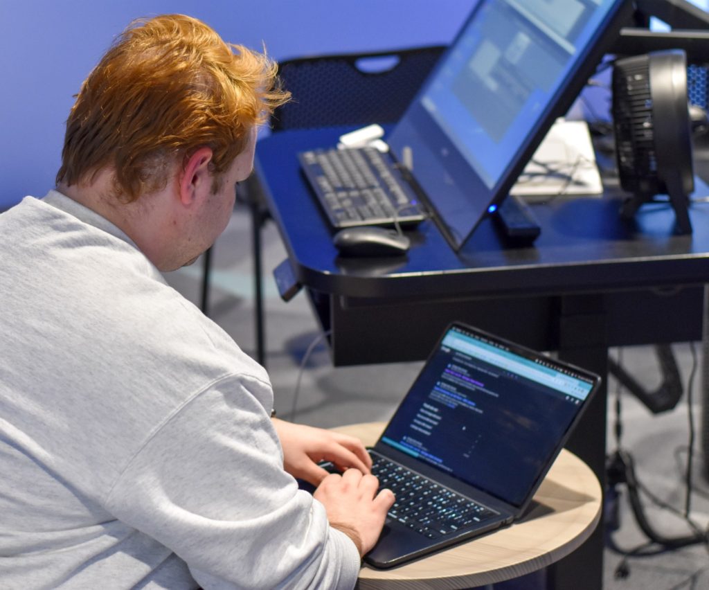 A person with red hair wearing a grey sweater works on their laptop.