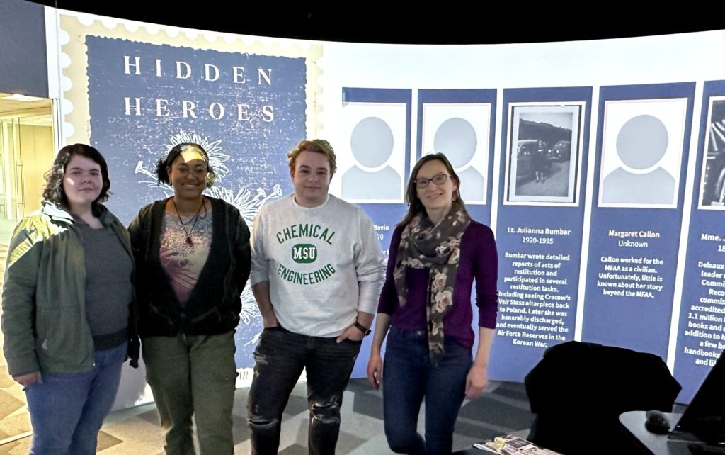 A picture of four people in a room. They are posing in front of a wall with images projected onto it. 