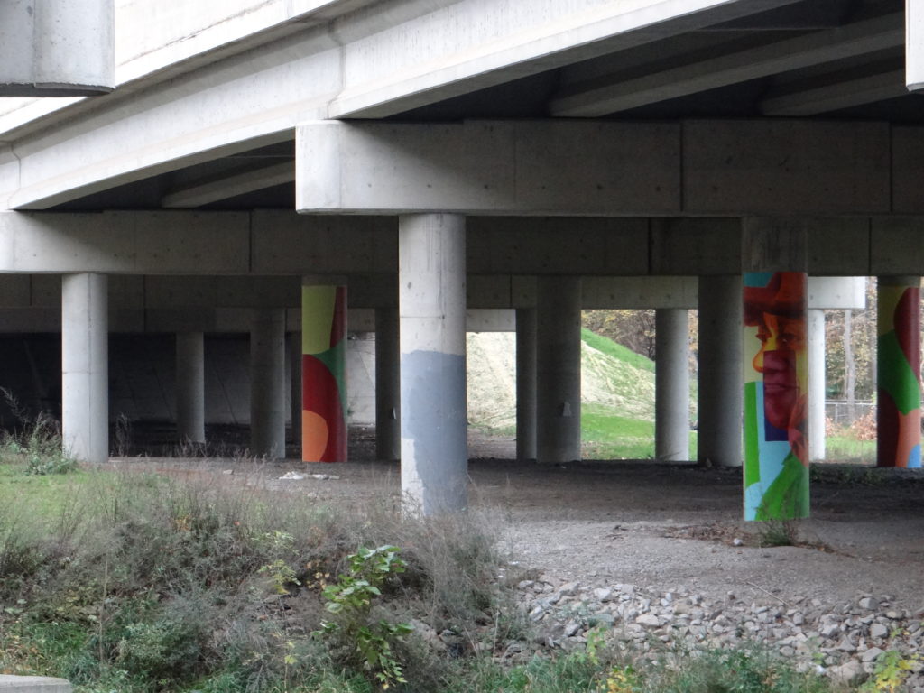 Underside of the I-496 bridge, supported by several concrete columns with colorful murals. 