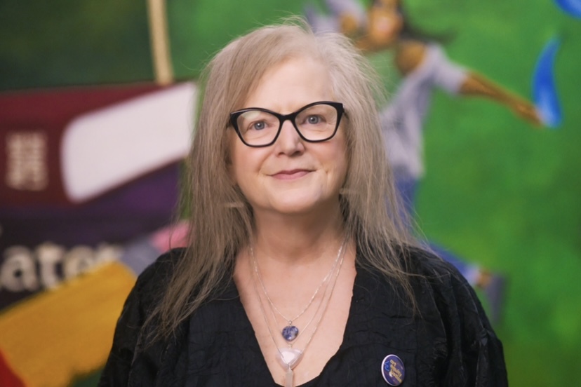 A picture of a woman in black with black glasses in front of a multicolored background.