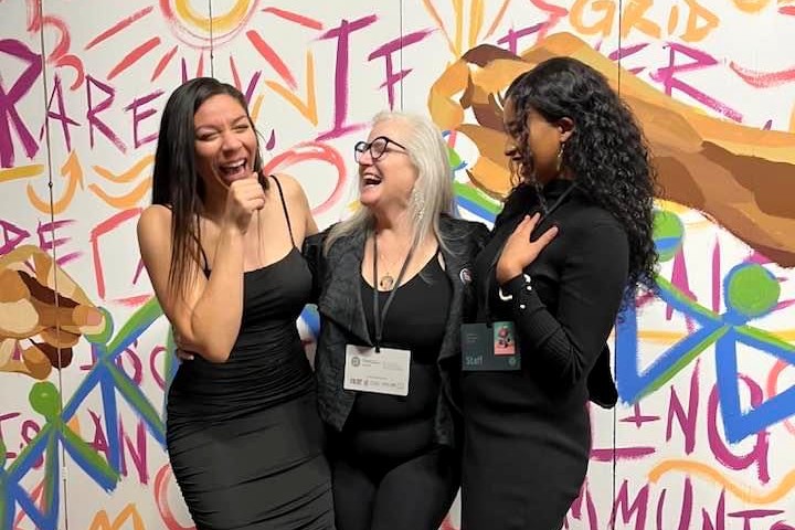 A picture of three women dressed in back laughing in front of a colorful background.