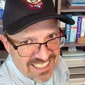man wearing baseball hat and glasses, smiling in front of a bookshelf
