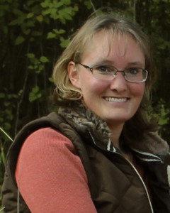 Imari, a white woman with short light brown hair and glasses, is smiling in front of a leafy background.