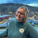 A smiling person wearing glasses and a green “Ventura Beach, California” sweatshirt. They are standing on a balcony overlooking a quaint town and a hill with trees.