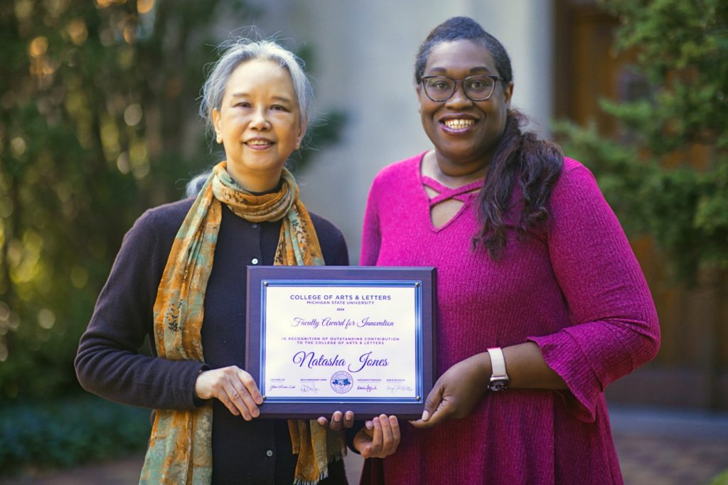 Two women stand outside, smiling, holding a certificate. The woman on the left is dressed in a dark sweater with a patterned scarf, and the woman on the right wears a bright magenta top. Behind them is green foliage and a brown door. 