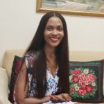 A brown skinned, smiling lady with long hair that has middle parting, wearing a floral dress, sitting on a cream white couch with green and red cushions, facing the camera in front of her.