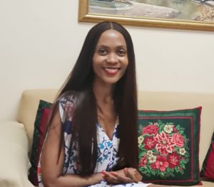 A brown skinned, smiling lady with long hair that has middle parting, wearing a floral dress, sitting on a cream white couch with green and red cushions, facing the camera in front of her.