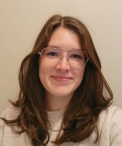 Sydney, a white woman with long brown hair and glasses, smiling.