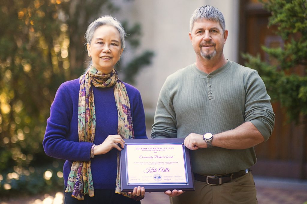 A woman in a purple sweater and patterned scarf and a man in a grey-green long-sleeve shirt stand outside, holding an award. Behind them is green foliage and a brown door. 